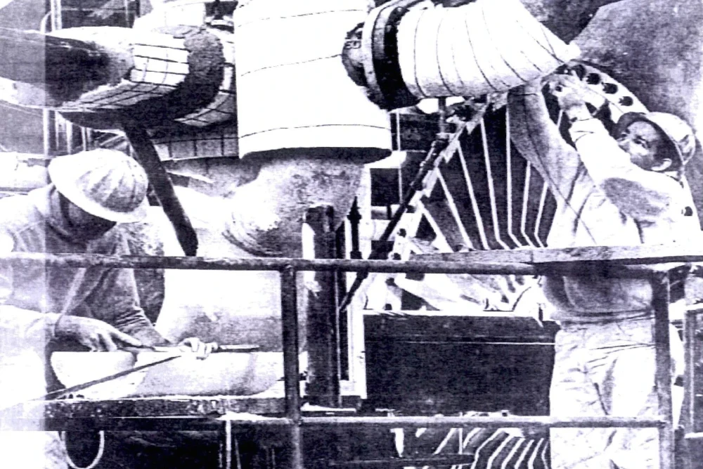 Tradesman (standing) works on insulating pipe that is a 90-degree bend. Tradesman (sitting) cuts pipe insulation with a keyhole saw. Although these insulating parts contained asbestos, neither tradesman are seen wearing any masks or respiratory protection.