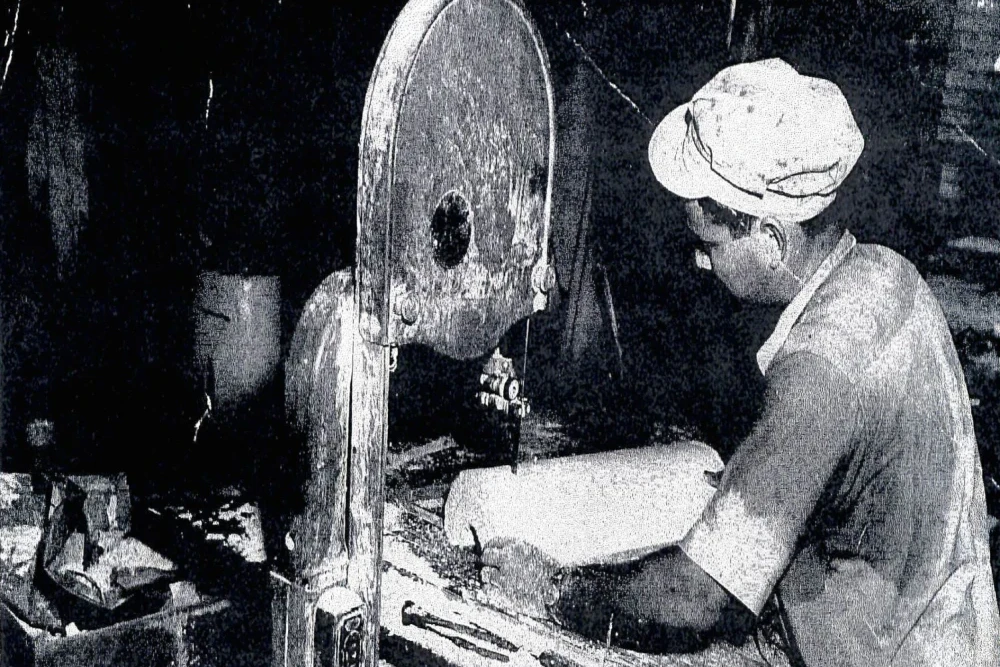 An industrial worker uses a band saw to cut pieces of pipe insulation. The white dust (which contains microscopic asbestos fibers) covers his hand, work station, and electrical outlet.